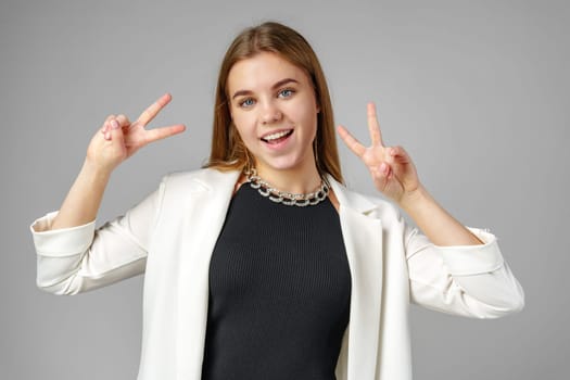 Young Woman in Stylish Attire Giving Peace Signs With Both Hands Against a Grey Background close up