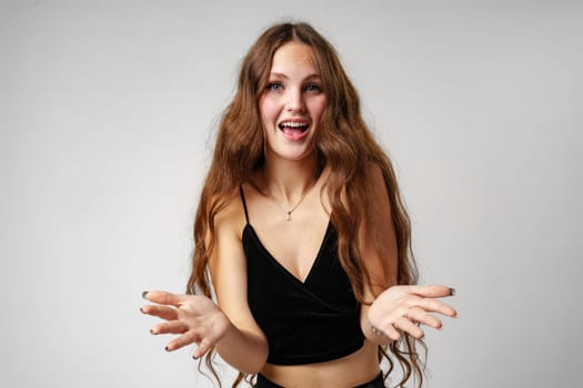 A young woman with long, wavy hair smiles broadly and gestures animatedly with her hands, expressing excitement and happiness. She is wearing a black V-neck top and stands against a simple, uncluttered backdrop that accentuates her lively demeanor.