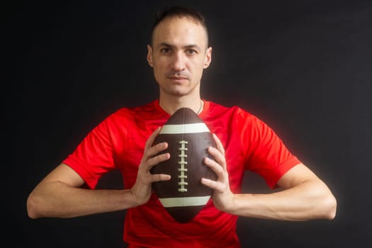 Young man dressed in casual street clothes holds a football, ready to throw a pass, isolated on black background. High quality photo