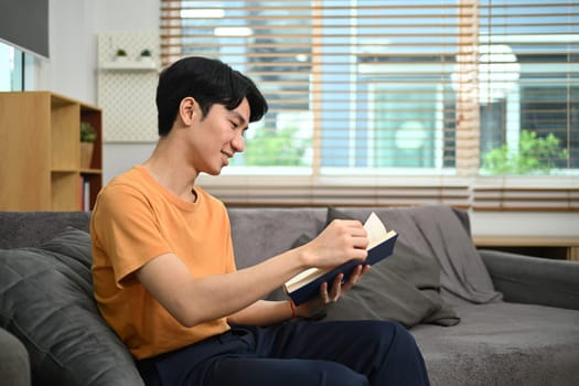 Smiling man relaxing on couch in living room and reading book. Hobby, leisure and knowledge concept.
