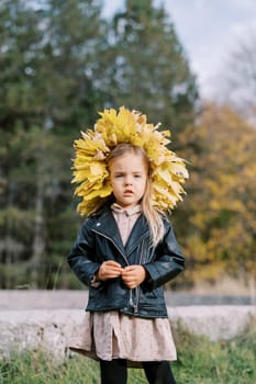 Little serious girl in a wreath of yellow leaves stands on the lawn. High quality photo