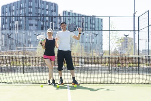 Portrait of positive young woman and adult man standing on padel tennis court, holding racket and ball, smiling. High quality photo