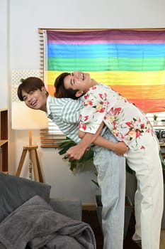 Cheerful gay male couple giving piggyback ride for each other laugh joyfully in living room.