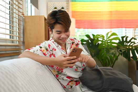 Relaxed gay young man using mobile phone while sitting in living room with rainbow flag.