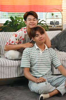 Portrait of beautiful gay couple smiling and looking at camera while sitting on couch at home.