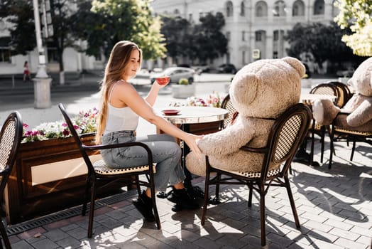 A woman sits cafe with a teddy bear next to her. The scene is set in a city with several chairs and tables around her. The woman is enjoying her time at the outdoor cafe