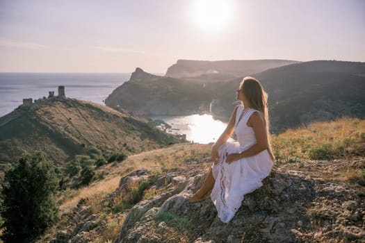 A woman in a white dress sits on a rock overlooking a body of water. The scene is serene and peaceful, with the woman enjoying the view and the calmness of the surroundings