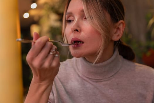 A woman is eating with a fork and spoon. She is wearing a gray shirt and has her hair in a ponytail