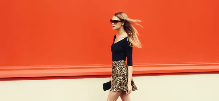 Beautiful stylish blonde young woman in leopard print skirt with black handbag clutch walking along on city street