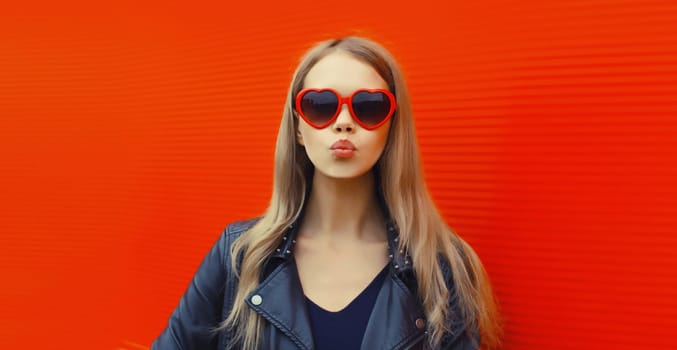 Portrait of beautiful stylish blonde young woman model blowing kiss in red heart shaped sunglasses, black rock leather jacket on city street