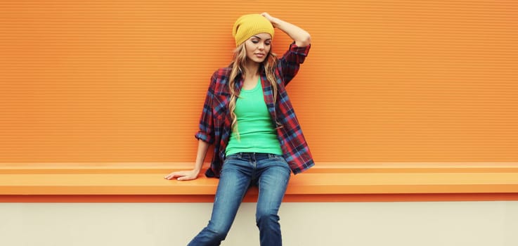 Beautiful young blonde woman posing in yellow hat, casual shirt on colorful orange background in the city
