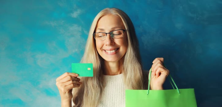 Portrait of happy smiling caucasian middle aged woman holding plastic credit bank card with shopping bags on blue background