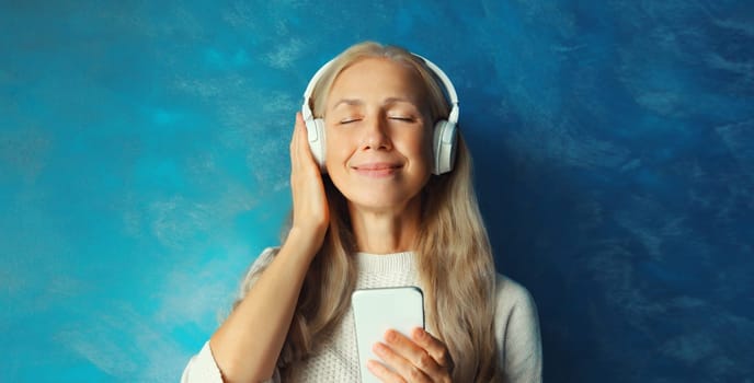 Portrait of happy smiling caucasian middle aged woman listening to music in headphones with smartphone on blue background