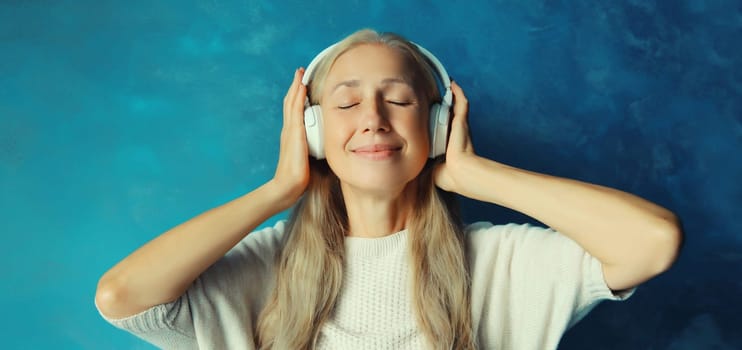 Portrait of happy relaxed middle aged woman listening to music in wireless headphones on blue background
