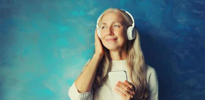 Portrait of happy smiling caucasian middle aged woman listening to music in headphones with smartphone on blue background