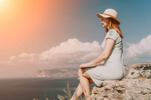 Digital nomad, woman in the hat, a business woman with a laptop sits on the rocks by the sea during sunset, makes a business transaction online from a distance. Freelance, remote work on vacation.