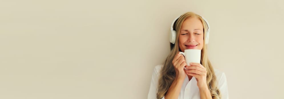 Portrait of happy relaxed middle aged woman listening to music drinking mug of coffee in headphones on white background, blank copy space