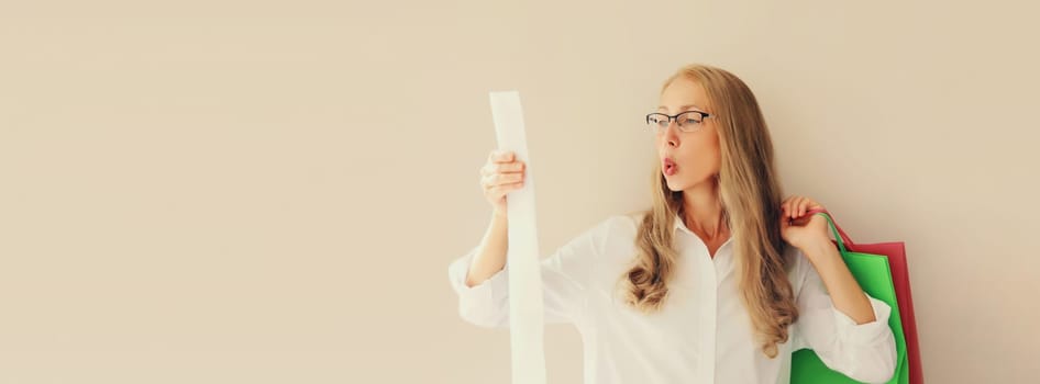 Shocked and surprised caucasian mature woman in eyeglasses looks amazed at the high price tag on long paper receipt holding shopping bag, copy space background