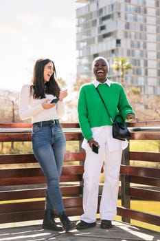 two young female friends laughing happy while having fun carefree using their mobile phones in a city park, concept of diversity and modern lifestyle, copy space for text