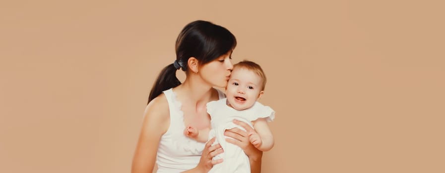 Happy young mother holding and kissing baby on brown studio background