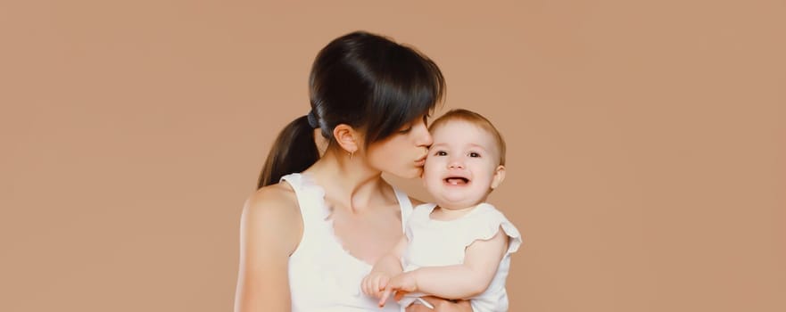 Happy young mother holding and kissing baby on brown studio background