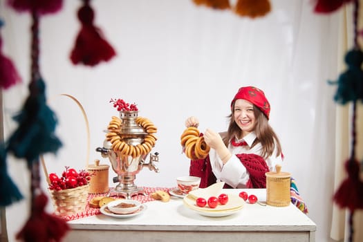 A fashionable modern girl in stylized folk clothes at table with a samovar, bagels and tea for the Orthodox holiday of Maslenitsa and Easter. Funny photo shoot for a young woman