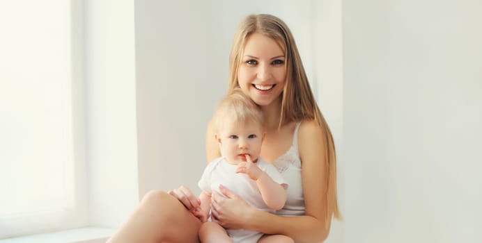 Happy smiling young mother playing with cute baby sitting together in white room at home