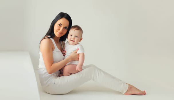 Happy young mother holding her cute baby sitting together in white room at home