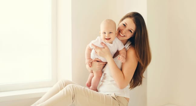 Happy smiling young mother playing with cute baby sitting together in white room at home