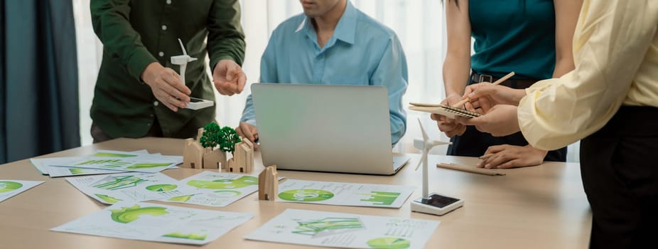 Cropped image of skilled businessman presented eco-friendly house project to general manager at meeting table with wooden block, windmill model and environmental document scatter around. Delineation.