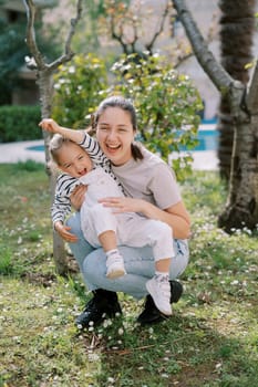 Laughing mother holds laughing little girl on her lap while squatting in garden. High quality photo