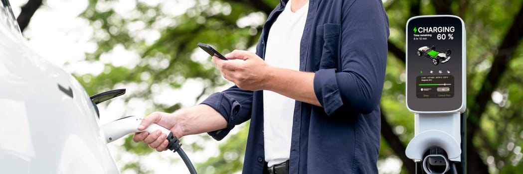 Man using smartphone online banking application to pay for electric car battery charging from EV charging station during vacation road trip at national park or summer forest. Panorama Exalt