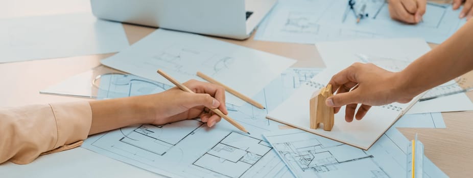 Professional architect hand draws a blueprint on table with architectural document and wooden block scatter around at office. Design and Planing concept. Focus on hand. Closeup. Delineation.