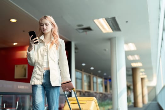 Young female traveler with suitcase using smartphone at airport. Modern travel, casual outfit, waiting for flight.