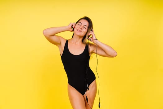 Caucasian Woman in Black Bodysuit Listening Music in Headphones On Yellow Background.