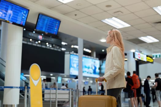Female traveler with suitcase checking flight information at airport. Modern travel concept, waiting for departure, casual outfit.