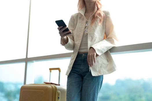 Young woman with suitcase using smartphone at airport terminal. Traveler waiting for flight, casual outfit..