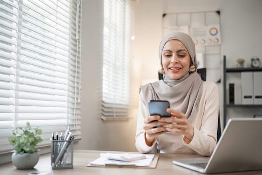 Smiling Muslim woman using smartphone at office desk. Professional workspace, casual attire.