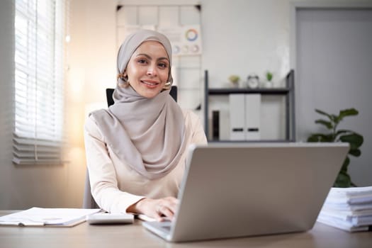 Smiling Muslim woman working on laptop at office desk. Professional workspace, casual attire.
