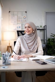 Smiling Muslim woman writing notes at office desk. Professional workspace, casual attire.