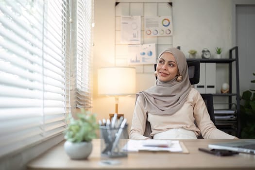 Smiling Muslim woman sitting at office desk. Professional workspace, casual attire.