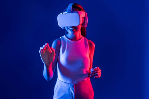 Smart female standing with surrounded by cyberpunk neon light wear VR headset connecting metaverse, futuristic cyberspace community technology. Woman using hand touching virtual object. Hallucination.