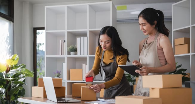 Two Asian women packing boxes and using a tablet in a home office. Concept of small business and e-commerce..