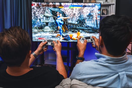 Friend gamers playing video game of battle martial arts fighter on TV using joysticks position of backside background. Comfy living neon light at home place with cheerful fighting winner. Sellable.