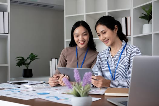 Two Asian businesswomen discussing work using a tablet in a modern office. Concept of teamwork and professional collaboration.