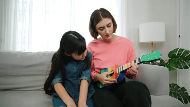 Happy girl playing ukulele while caucasian mom teaching and explain about acoustic music at home. Cute child learning about instrument. Happy caucasian mother and girl spend time together. Pedagogy.