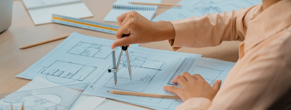 Professional architect drawing blueprint by using divider on the table with stationary and architectural document scatter around at architectural office. Closeup. Focus on hand. Delineation.