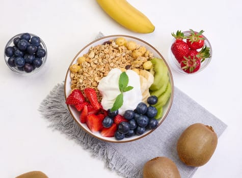 Granola with strawberries, kiwi, banana and blueberries in a round plate on a white table. Healthy and tasty food, top view