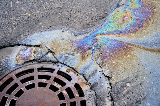A puddle of motor oil and gasoline on the asphalt road seeps into a nearby sewer grate