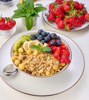 Granola with strawberries, kiwi, banana and blueberries in a round plate on a white table. Healthy and tasty food, top view